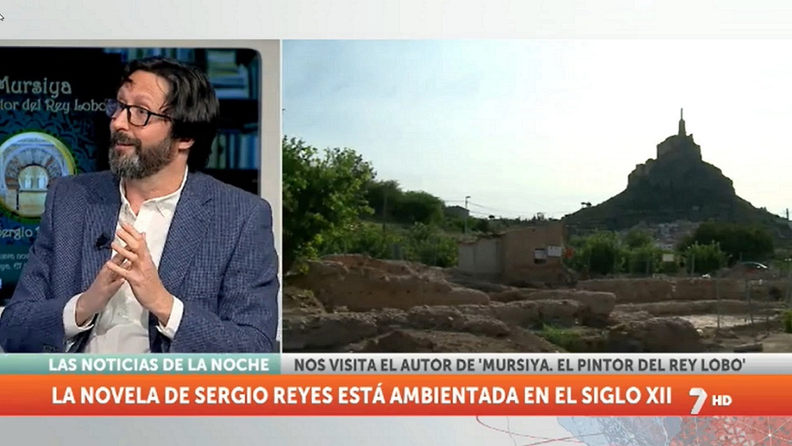 Sergio Reyes en 7TV Región de Murcia, con imágenes de los yacimientos de Monteagudo y el castillo al fondo, hablando de "Mursiya. El pintor del Rey Lobo" en las noticias de la noche de Luis Alcázar