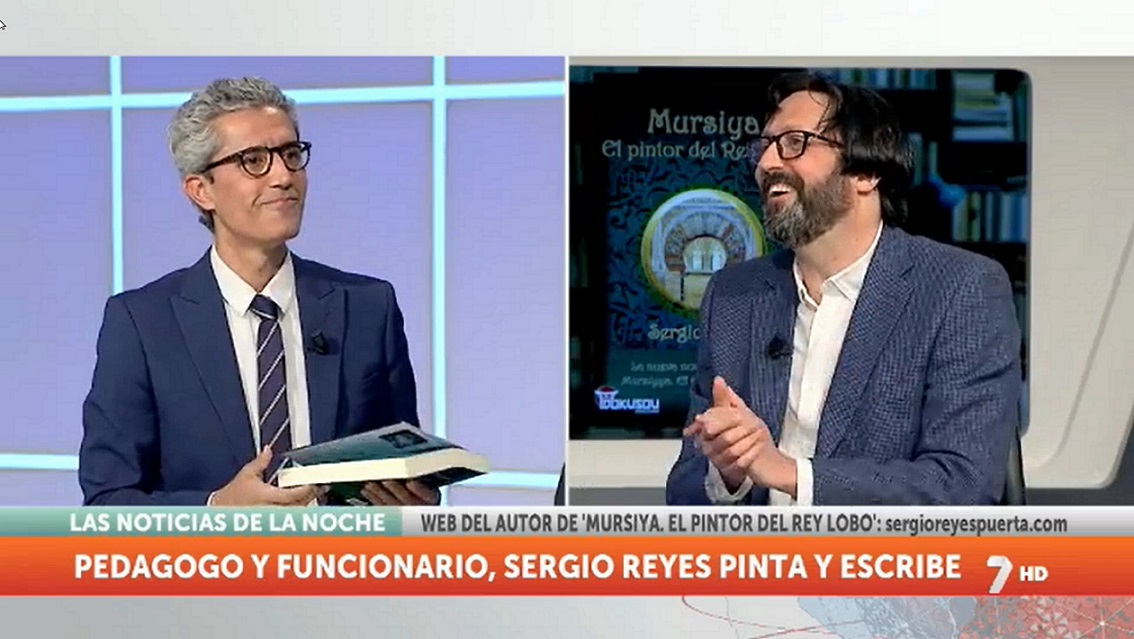 Luis Alcázar con un ejemplar de "Mursiya. El pintor del Rey Lobo" escucha al autor en las noticias de la noche de la 7 TV Región de Murcia